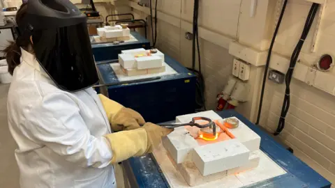 Kevin Church, BBC A scientist demonstrates how nuclear waste can be 'baked' into solid materials like ceramic by making ceramic in a the laboratory. The researcher wears a white coat and protective helmet as she uses gloves and tongs to handle a glowing-hot piece of ceramic