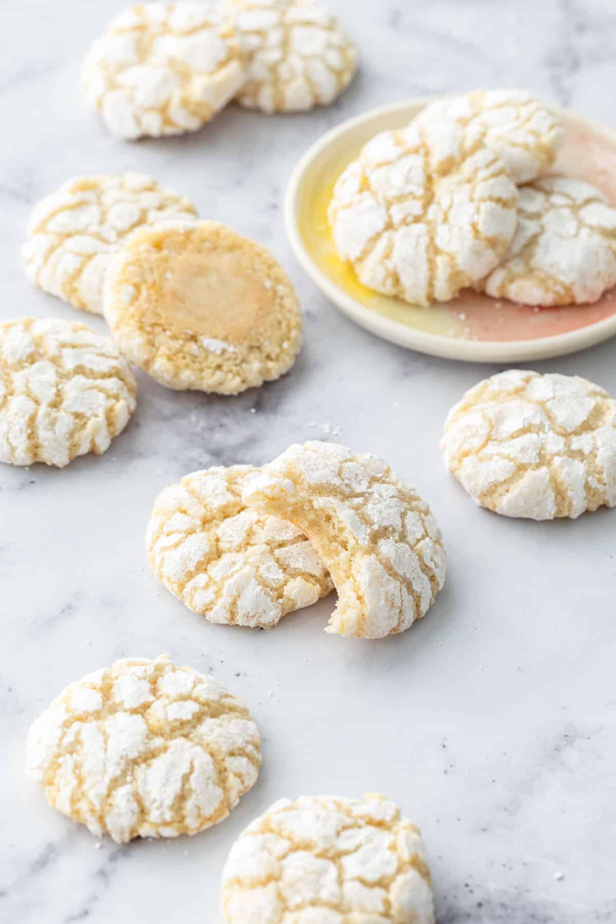 Chewy Lemon Almond Crinkle Cookies scattered on a marble background with a few cookies on a small cookie plate.