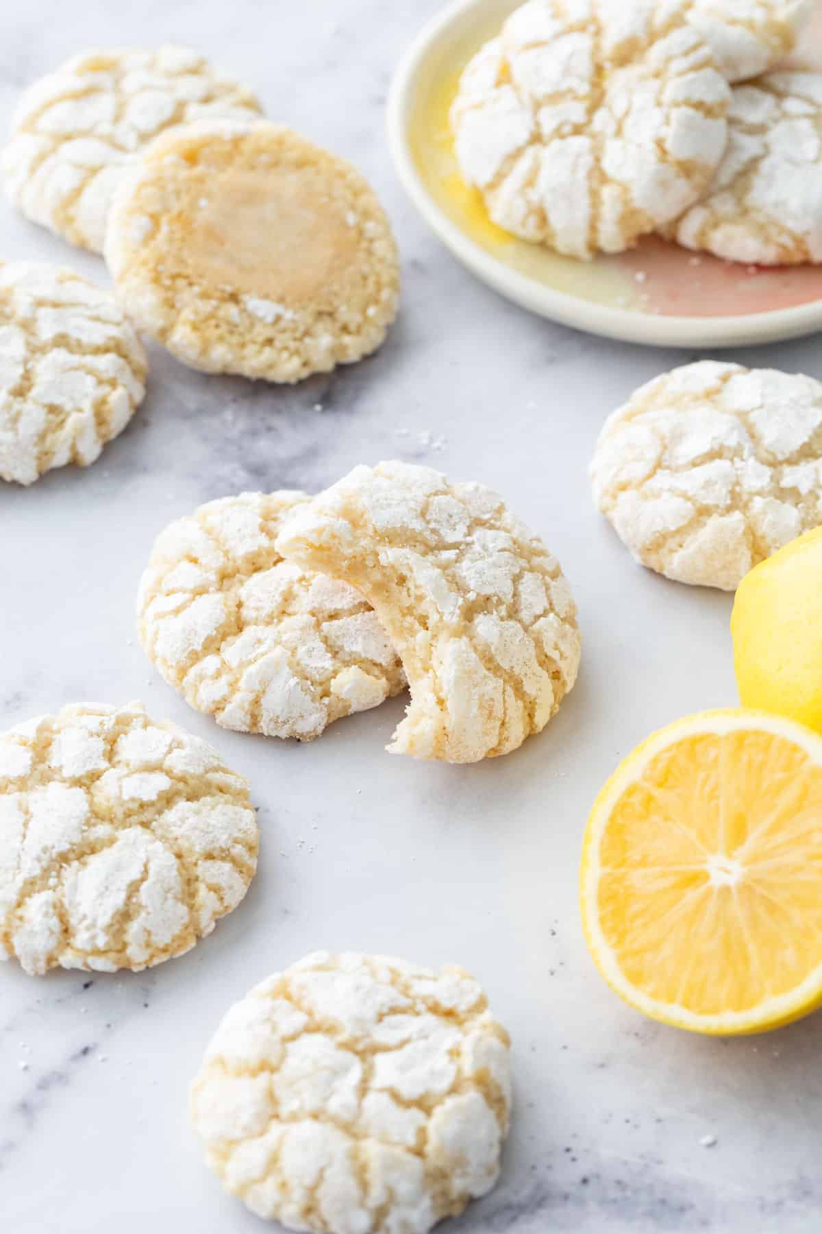 Chewy Lemon Almond Crinkle Cookies scattered on a marble background with a few cookies on a small cookie plate and a cut lemon in the foreground.