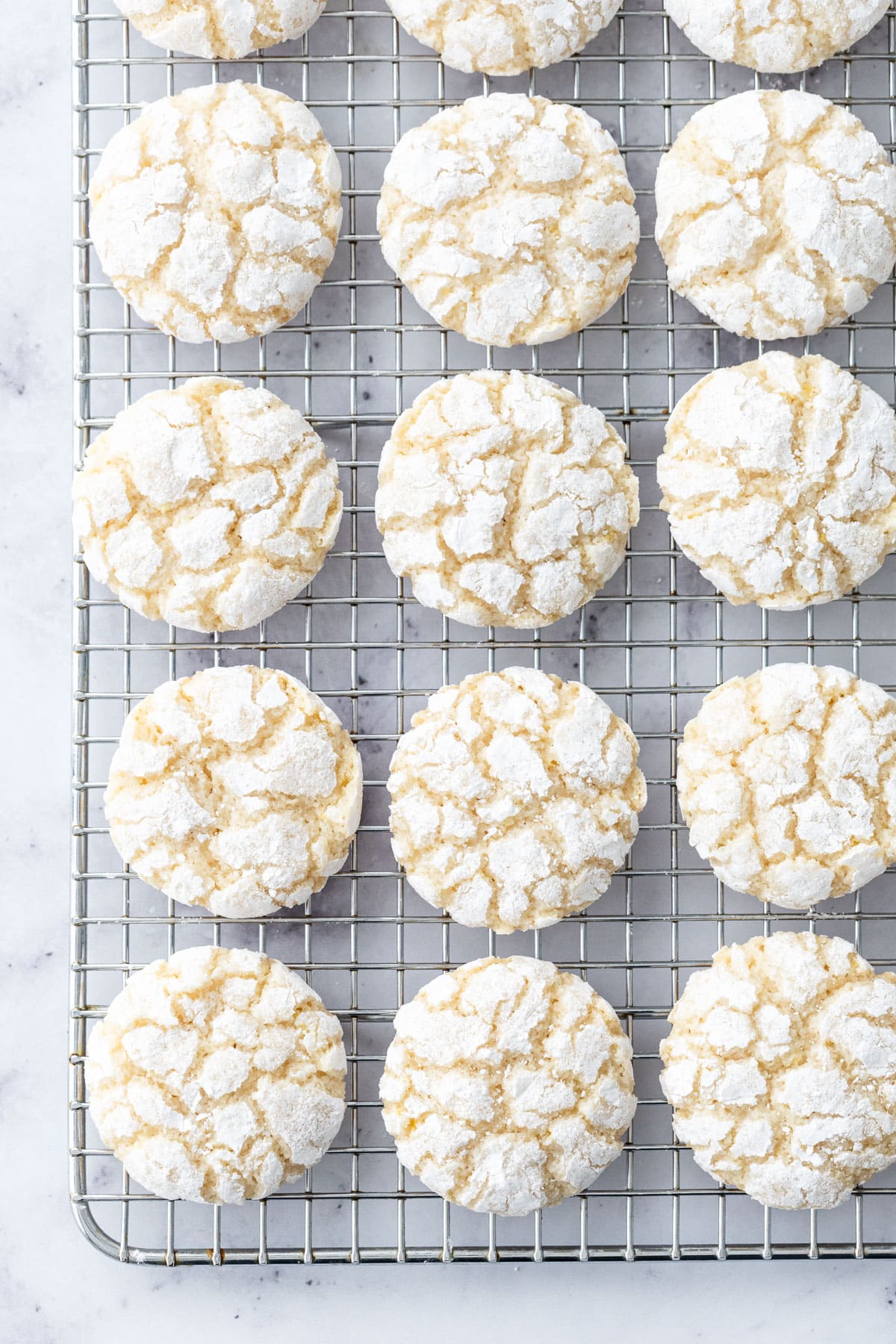 Overhead, neat rows of Chewy Lemon Almond Crinkle Cookies on a wire cooling rack sitting on a marble surface.