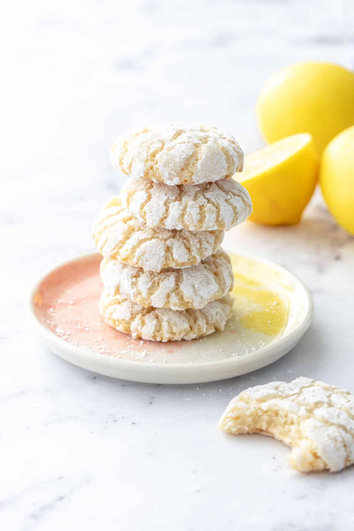 Stack of Chewy Lemon Almond Crinkle Cookies on a pink and yellow cookie plate, with lemons out of focus in the background.