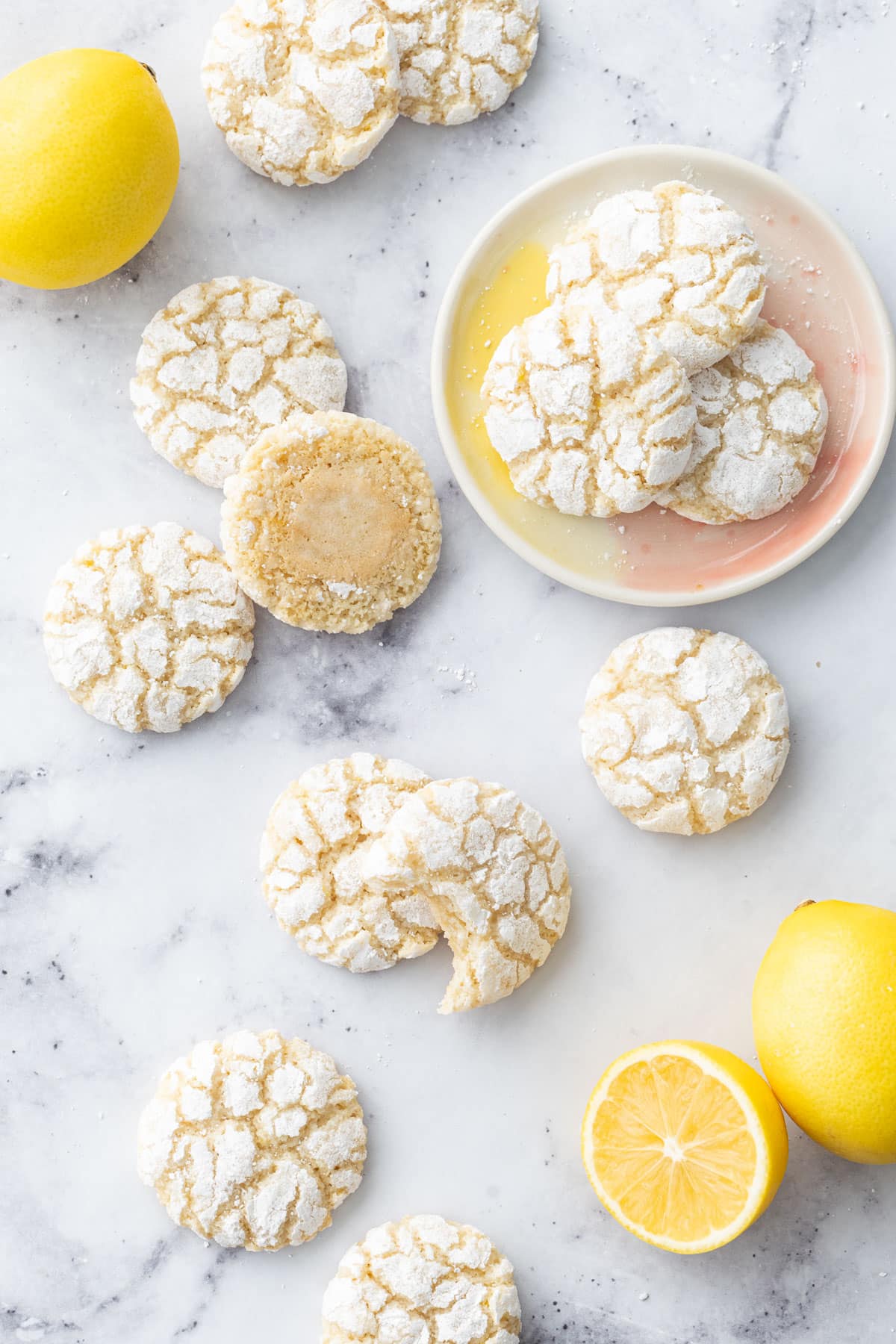 Overhead, scattered Chewy Lemon Almond Crinkle Cookies on a marble surface with a few fresh lemons and three cookies on a small ceramic plate.