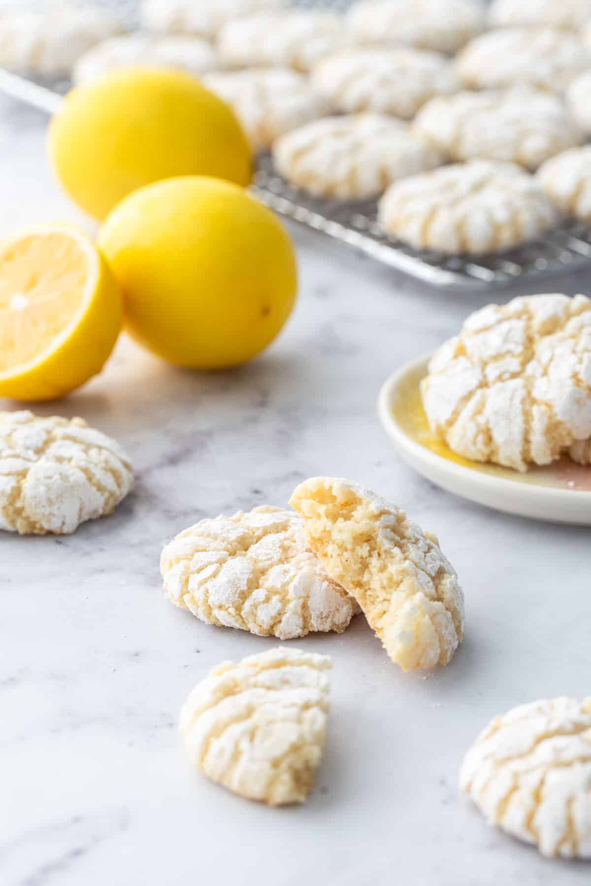 Chewy Lemon Almond Crinkle Cookies on a marble background, more cookies on a wire cooling rack and fresh lemons out of focus in the background.