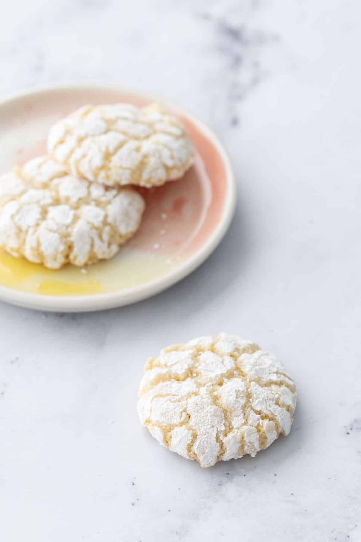 Two Chewy Lemon Almond Crinkle Cookies on a pink and yellow plate, one cookie sitting in front on the marble countertop surface.