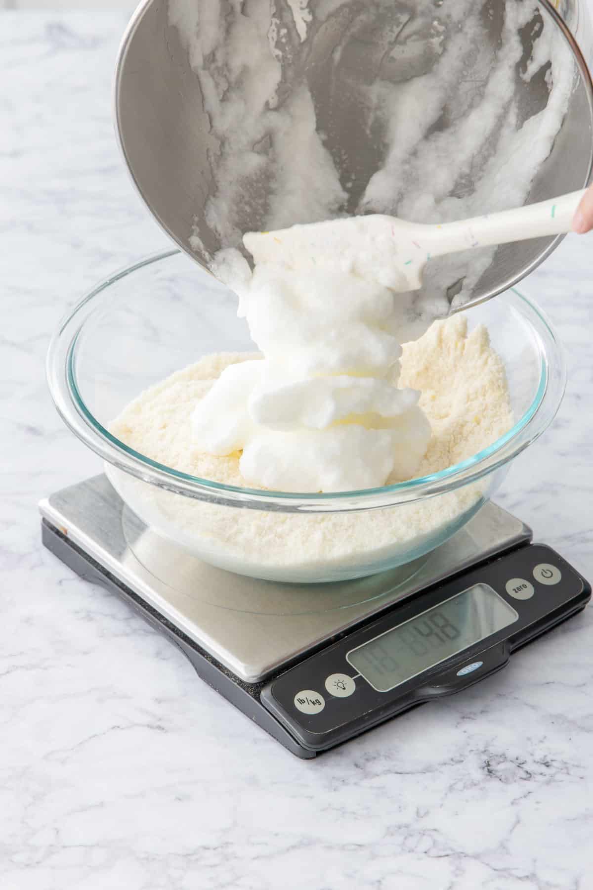 Scooping beaten egg whites into a mixing bowl along with almond flour and sugar.