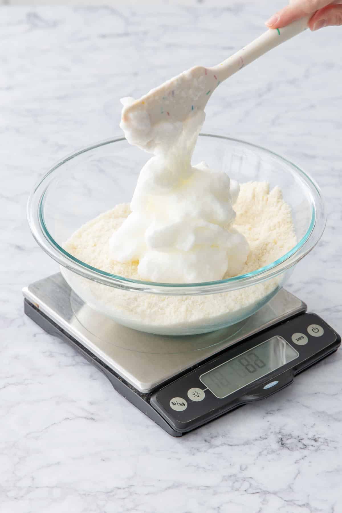 Scooping beaten egg whites into a mixing bowl along with almond flour and sugar.