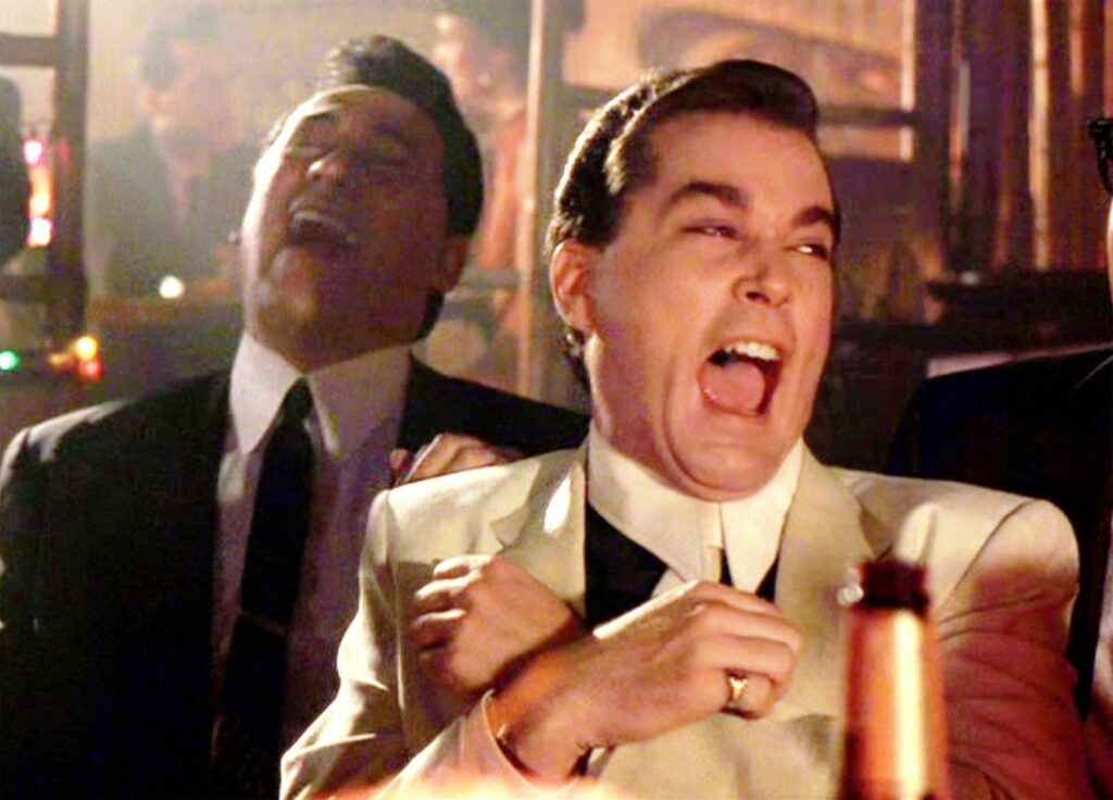 Two men in suits are sitting in a dimly lit bar, laughing heartily. The man in front is wearing a white suit and has his arms crossed in front of him, while the man behind him, also in a suit, leans back laughing. A beer bottle is visible.