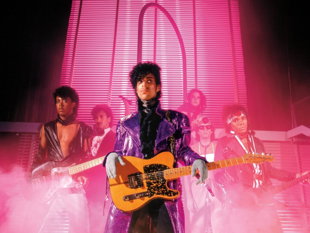 A band stands on stage in dramatic pink lighting and fog. The lead performer in front wears a shiny purple coat and holds a guitar, with other band members behind him playing instruments.